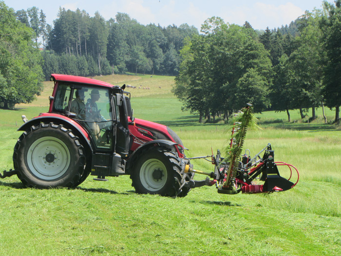  Das Foto zeigt einen roten Traktor auf einer landwirtschaftlich genutzten Grünfläche. In dem Zugfahrzeug sitzen zwei Menschen. Daran vorgespannt ist eine Mähmaschine mit Schneidwerk in Form eines Balkens. Der Fahrer mäht damit das Grünland. Im Hintergrund befinden sich mehrere Baumreihen und ein Waldsaum. Dazwischen sind vereinzelt Schafe auf einer Weidefläche zu erkennen.